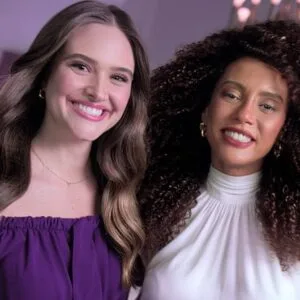 Atriz Juliana Paiva à esquerda, com blusa roxa, e atriz Taís Araújo, à esquerda, com busa branca, ambas sorrindo e com cabelos esvoaçantes.
